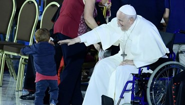 البابا فرنسيس جالسًا على كرسي متحرك يلاعب طفلا خلال لقاء عام ضمن مهرجان العائلات في قاعة بول السادس في الفاتيكان (22 حزيران 2022، أ ف ب). 