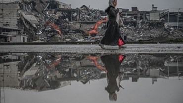 امرأة تمشي في شارع غمرته المياه بينما يعمل حفارون على تنظيف أنقاض أبنية منهارة في أنطاكيا بعد 11 شهرا من الزلزال (12 ك2 2024، أ ف ب).
